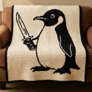 May include: A rectangular crochet blanket featuring a large, black and cream-coloured graphic of a penguin holding a knife. The design is rendered in a pixelated style with a solid black border framing the cream-coloured background of the throw.