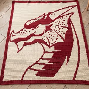 May include: A crocheted blanket featuring a dragon's head design. The dragon is rendered in red against a cream-colored background. The blanket has a red border. The design is a pixelated style, giving it a unique, handcrafted appearance.