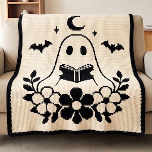 May include: A cream-colored crochet blanket featuring a black graphic design of a ghost reading a book. The ghost is surrounded by flowers, two bats, stars, and a crescent moon. The blanket has a solid black border along all four edges.