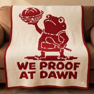 Peut inclure: Une couverture en crochet couleur crème avec un motif rouge foncé représentant une grenouille portant un tablier, tenant un pain dans un panier et une cuillère en bois. Le texte WE PROOF AT DAWN est inscrit en lettres capitales rouge foncé.