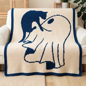 Könnte beinhalten: Eine cremefarbene Häkeldecke mit einem großen, dunkelblauen Grafikdesign, das eine Katze im Zusammenspiel mit einem Geist zeigt. Die Decke hat einen durchgehenden dunkelblauen Rand an allen vier Seiten und eine sichtbare, gitterartige Häkelstruktur.