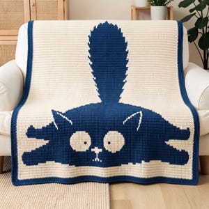 Peut inclure: Une couverture en crochet rectangulaire présentant un fond crème avec un grand motif bleu foncé représentant un chat allongé. Le chat a une longue queue dressée, de grands yeux circulaires et un petit nez. La couverture a une bordure bleu foncé.