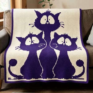 May include: A rectangular crochet blanket featuring a cream-colored background with a dark purple graphic pattern of three stylized cats. The cats have large, wide eyes and long tails that curve outward at the bottom edges of the textile piece.
