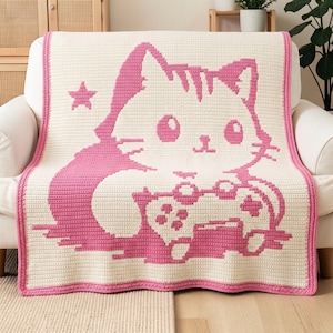 Puede incluir: Una manta de ganchillo de color crema con un gran gráfico rosa de un gato sosteniendo un mando de videojuegos. El diseño incluye una pequeña estrella rosa a la izquierda del gato, y toda la manta está rematada con un borde rosa.