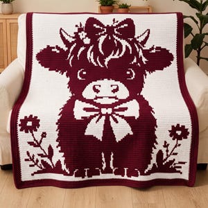 May include: A rectangular crochet blanket featuring a two-tone maroon and white design. The centre displays a large highland cow with a bow on its head and a bow around its neck, flanked by two small flowers at the bottom corners.