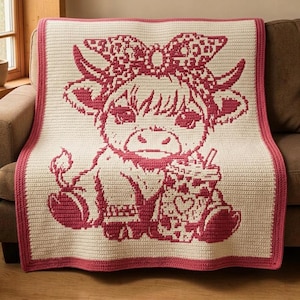May include: A cream-colored crochet blanket featuring a dark pink graphic of a highland cow wearing a bow and holding a drink. The blanket has a solid dark pink border around the entire rectangular edge, creating a distinct frame for the design.