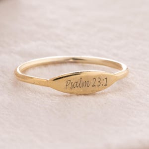 Może przedstawiać: Cienki złoty pierścionek z małą, płaską, owalną płytką środkową. Płytka jest wygrawerowana tekstem Psalm 23:1 kursywą. Obrączka jest smukła i okrągła, zachowując spójne metaliczne złote wykończenie na całej powierzchni elementu.