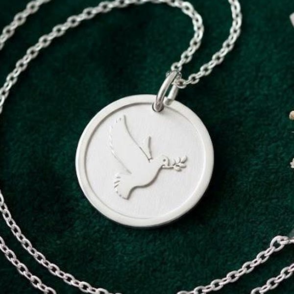 May include: A silver-toned circular pendant necklace featuring an embossed dove carrying an olive branch in its beak. The pendant is suspended from a delicate matching silver-toned chain with a circular spring ring clasp, showcasing a clean design.
