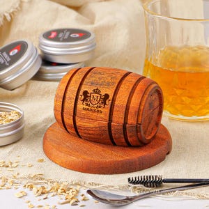 Puede incluir: Un ahumador de madera en forma de barril con detalles grabados, apoyado sobre una base redonda de madera. El barril presenta un escudo y el nombre "Madison". También hay varias latas de metal pequeñas con etiquetas y un vaso de líquido ámbar.