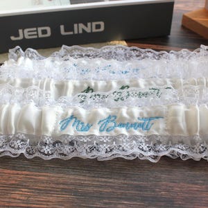 May include: White satin wedding garter with delicate lace trim. The garter features the embroidered name "Mrs. Bovitt" in light blue thread. A romantic accessory for a bride.