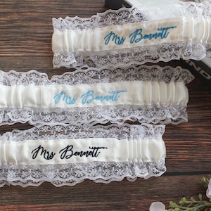 May include: Three white elastic garters with white lace trim. Each garter features the embroidered text "Mrs Bennett" in different thread colors: dark blue, light blue, and black. The garters are arranged horizontally on a dark wood surface.