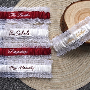 Può includere: Diverse giarrettiere da sposa in raso bianco e rosso con finiture in pizzo sono esposte. Alcune giarrettiere hanno nomi ricamati, come "The Smiths", "The Sakals", "Payday" e "Mrs. Hernandez". Una giarrettiera è su una fetta di legno, altre su una superficie intrecciata.