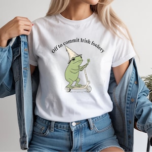 May include: White t-shirt featuring a cartoon frog riding a scooter, wearing a pointed hat and holding a four-leaf clover. The text "Off to commit Irish foolery" is printed above the frog.