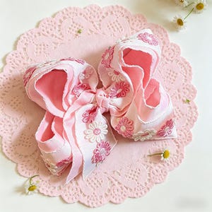 May include: A large, pink hair bow with white and pink floral embroidery. The bow is made of layered ribbons and sits on a pink, lace-edged doily. Small daisies are scattered around the bow.
