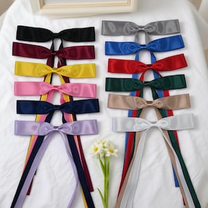 May include: A collection of satin ribbon bows in various colors, including black, gray, blue, red, green, tan, white, yellow, pink, and purple. Each bow has long, matching ribbons. The bows are arranged on a white surface.
