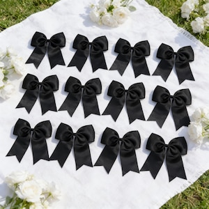 May include: A collection of black satin bows arranged on a white cloth, with white flowers in the background. The bows are evenly spaced and have long, pointed tails. The fabric has a slight sheen, and the bows appear to be handmade.