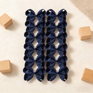 May include: A collection of navy blue hair bows arranged in rows. The bows are made of a textured fabric and have a classic bow shape. Wooden blocks are scattered around the bows.