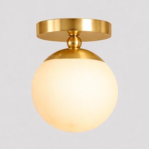 May include: A modern ceiling light fixture with a gold-colored metal base and a spherical, frosted glass shade. The light has a simple, elegant design, suitable for various interior styles. The fixture is designed to be mounted flush against the ceiling.