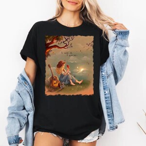 May include: A black short-sleeved t-shirt featuring a graphic illustration of a person with long blonde hair wearing a hat, sitting under a tree with a guitar. The text "I knew it. I knew you..." is printed above the figure in a sunset landscape.