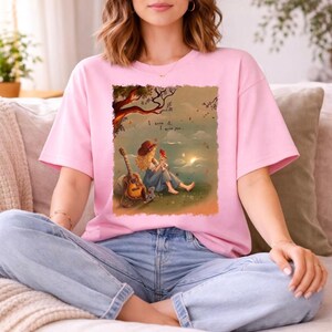 May include: A light pink t-shirt featuring a graphic illustration of a person sitting under a tree with a guitar and a small bird. The text "I knew it, I knew you..." is printed above the figure. The scene depicts a landscape with a body of water.