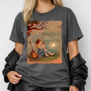 May include: A charcoal gray crew neck t-shirt featuring a graphic illustration of a person with blonde hair sitting on grass, holding a red bird, with an acoustic guitar nearby. The text reads, I knew it, I know you, under a tree branch.
