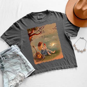 May include: A charcoal gray t-shirt featuring a graphic illustration of a person with blonde hair wearing a hat, sitting on the grass with a guitar and a small bird. The text "I knew it. I knew you..." is printed above the figure in a soft, warm-toned landscape.