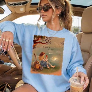 May include: A light blue crewneck sweatshirt featuring a rectangular graphic print of a person with curly hair sitting on grass next to an acoustic guitar, holding a small red bird. The text "I knew it, I knew you..." is printed above the figure.