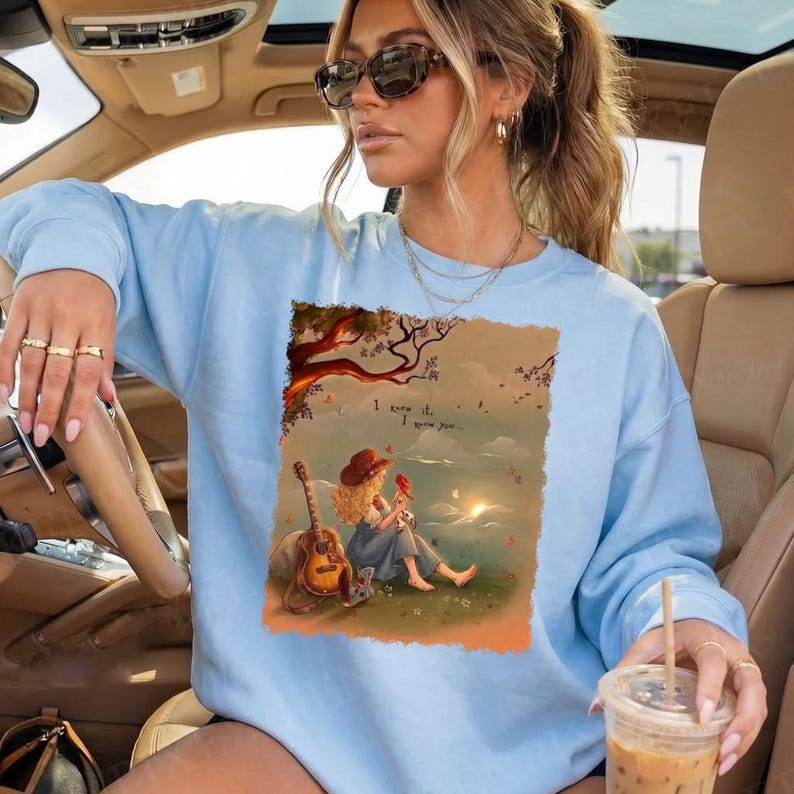 May include: A light blue crewneck sweatshirt featuring a rectangular graphic print of a person with curly hair sitting on grass next to an acoustic guitar, holding a small red bird. The text "I knew it, I knew you..." is printed above the figure.