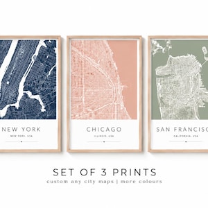 May include: A set of three framed wall art prints featuring stylized city maps of New York, Chicago, and San Francisco. Each print displays a detailed street grid in white against a solid background of navy blue, peach, or sage green.