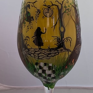 May include: A hand-painted wine glass with a whimsical Alice in Wonderland scene. The glass features a silhouette of Alice standing in a forest with a pocket watch hanging from a tree branch. The background is a yellow-gold color with green and black details.