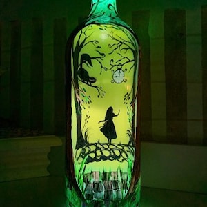 May include: A glass bottle with a green glow, painted with a silhouette of Alice in Wonderland standing in a forest with a rabbit and a pocket watch. The bottle is lit from the bottom, creating a magical effect.