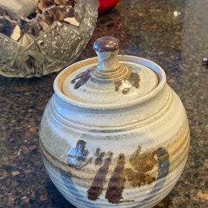 May include: A white ceramic jar with a lid, featuring brown and blue abstract designs. The jar has a rounded shape and a smooth, glazed finish.