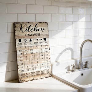 以下が含まれることがあります： 「Kitchen」の文字がエレガントな筆記体で書かれた、素朴な木製のキッチン変換サイン。このサインには、カップ、オンス、グラム、大さじ、小さじ、ミリリットルの換算表と温度換算が表示されています。
