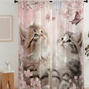 Peut inclure: Une paire de rideaux de fenêtre avec un motif imprimé représentant deux chatons duveteux assis parmi des fleurs de cerisier roses et des papillons. Les chatons ont un pelage brun clair et gris sur un fond à motif floral rose pâle.