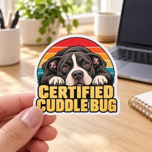 May include: A die-cut sticker featuring a black and white puppy resting its chin on its paws. The background shows a sunset with horizontal stripes in red, orange, and teal. The text CERTIFIED CUDDLE BUG is printed in bold yellow letters.
