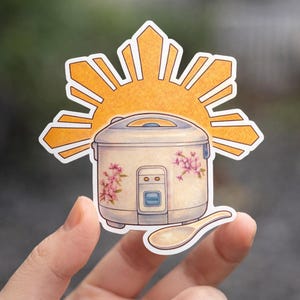 May include: A sticker featuring a rice cooker with floral accents and a wooden spoon. The rice cooker is set against a stylized orange sunburst design. The sticker is held up by a hand.