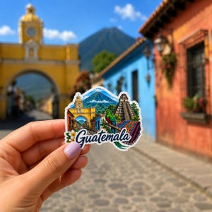 May include: A colorful Guatemala sticker featuring a vibrant illustration of a cityscape, a volcano, and a pyramid. The sticker has the word "Guatemala" in a decorative font. The background shows a blurred street scene with colorful buildings.
