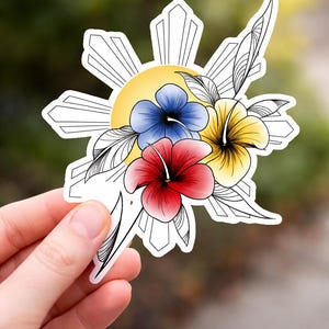 May include: A sticker featuring a yellow sunburst design with three colorful flowers: a blue, a yellow, and a red hibiscus. The flowers are surrounded by black and white leaves. The sticker is held in a hand.