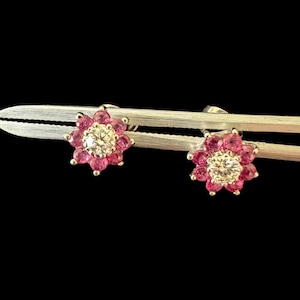 May include: A pair of stud earrings featuring a central round white diamond surrounded by a halo of eight small round pink rubies. The earrings are held by silver-toned metal tweezers against a solid black background, showcasing a floral design.