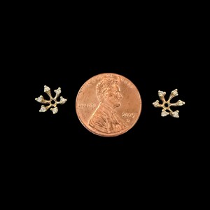 May include: A pair of small, gold-colored snowflake earrings with clear, sparkling accents. The earrings are displayed on a black background next to a U.S. penny for size comparison. The earrings have a delicate, open design.