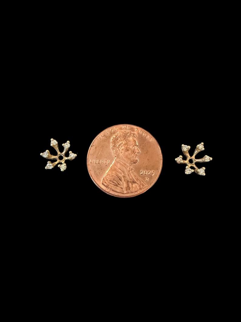 May include: A pair of small, gold-colored snowflake earrings with clear, sparkling accents. The earrings are displayed on a black background next to a U.S. penny for size comparison. The earrings have a delicate, open design.