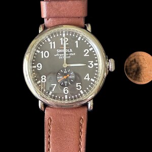 K&ouml;nnte beinhalten: Eine runde Armbanduhr mit dunkelgrauem Zifferblatt, silbernem Geh&auml;use und braunem Lederarmband. Das Zifferblatt zeigt wei&szlig;e Ziffern und Zeiger, mit einem kleineren Hilfszifferblatt. Das Armband ist gen&auml;ht.