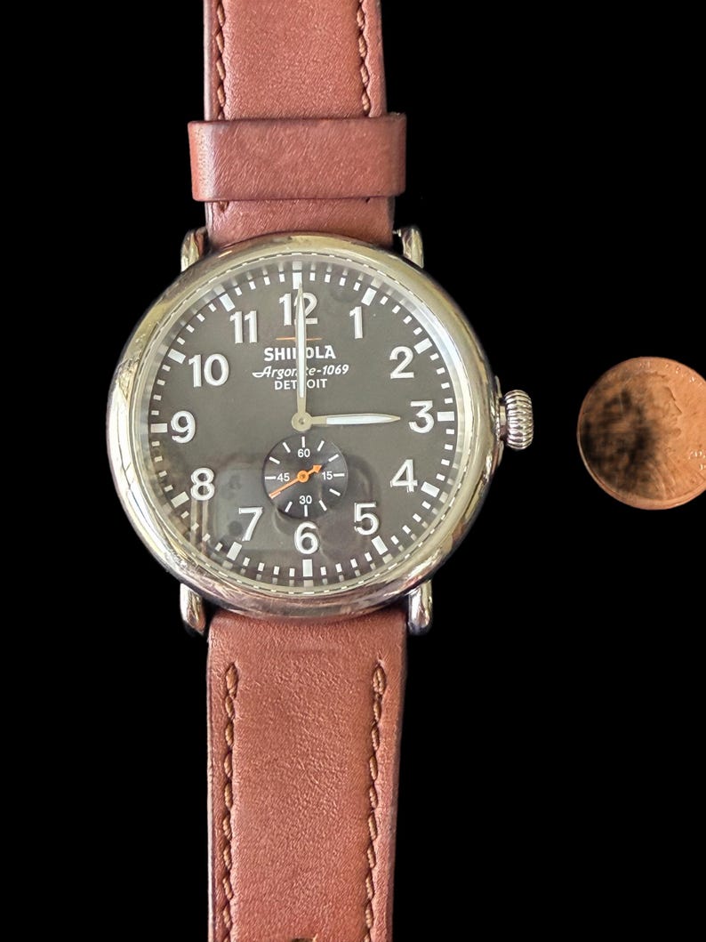 K&ouml;nnte beinhalten: Eine runde Armbanduhr mit dunkelgrauem Zifferblatt, silbernem Geh&auml;use und braunem Lederarmband. Das Zifferblatt zeigt wei&szlig;e Ziffern und Zeiger, mit einem kleineren Hilfszifferblatt. Das Armband ist gen&auml;ht.