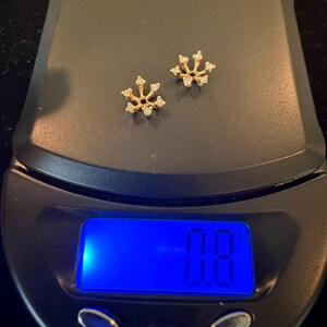 May include: Two gold-colored snowflake stud earrings with small, clear gemstones are displayed on a digital scale. The scale shows a weight of 7.8 grams. The scale has an on/off button, a mode button, and a tare button.