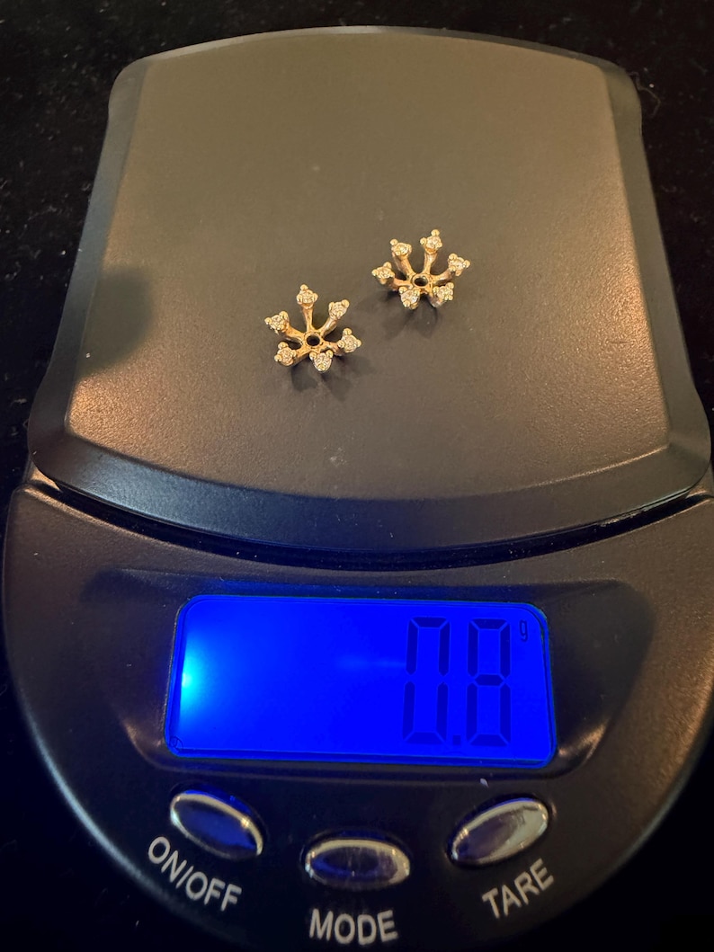 May include: Two gold-colored snowflake stud earrings with small, clear gemstones are displayed on a digital scale. The scale shows a weight of 7.8 grams. The scale has an on/off button, a mode button, and a tare button.
