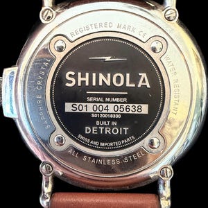 K&ouml;nnte beinhalten: Nahaufnahme der R&uuml;ckseite einer Shinola-Uhr mit einem braunen Lederarmband. Die Uhr hat ein Edelstahlgeh&auml;use mit dem Text "SHINOLA DETROIT" und "ALL STAINLESS STEEL". Die Uhr ist wasserdicht.