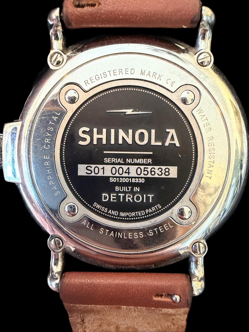 K&ouml;nnte beinhalten: Nahaufnahme der R&uuml;ckseite einer Shinola-Uhr mit einem braunen Lederarmband. Die Uhr hat ein Edelstahlgeh&auml;use mit dem Text "SHINOLA DETROIT" und "ALL STAINLESS STEEL". Die Uhr ist wasserdicht.