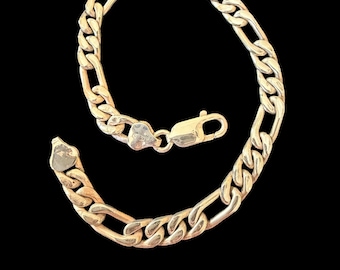 May include: A gold-toned Figaro chain bracelet featuring a series of flattened oval links interspersed with longer, elongated oval links. The bracelet is secured with a lobster claw clasp and includes a small, flat, heart-shaped metal tag.