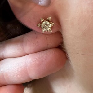 May include: A gold stud earring with a large, round, clear gemstone at the center, surrounded by smaller, clear gemstones in a starburst design. The earring is set in a gold metal setting.