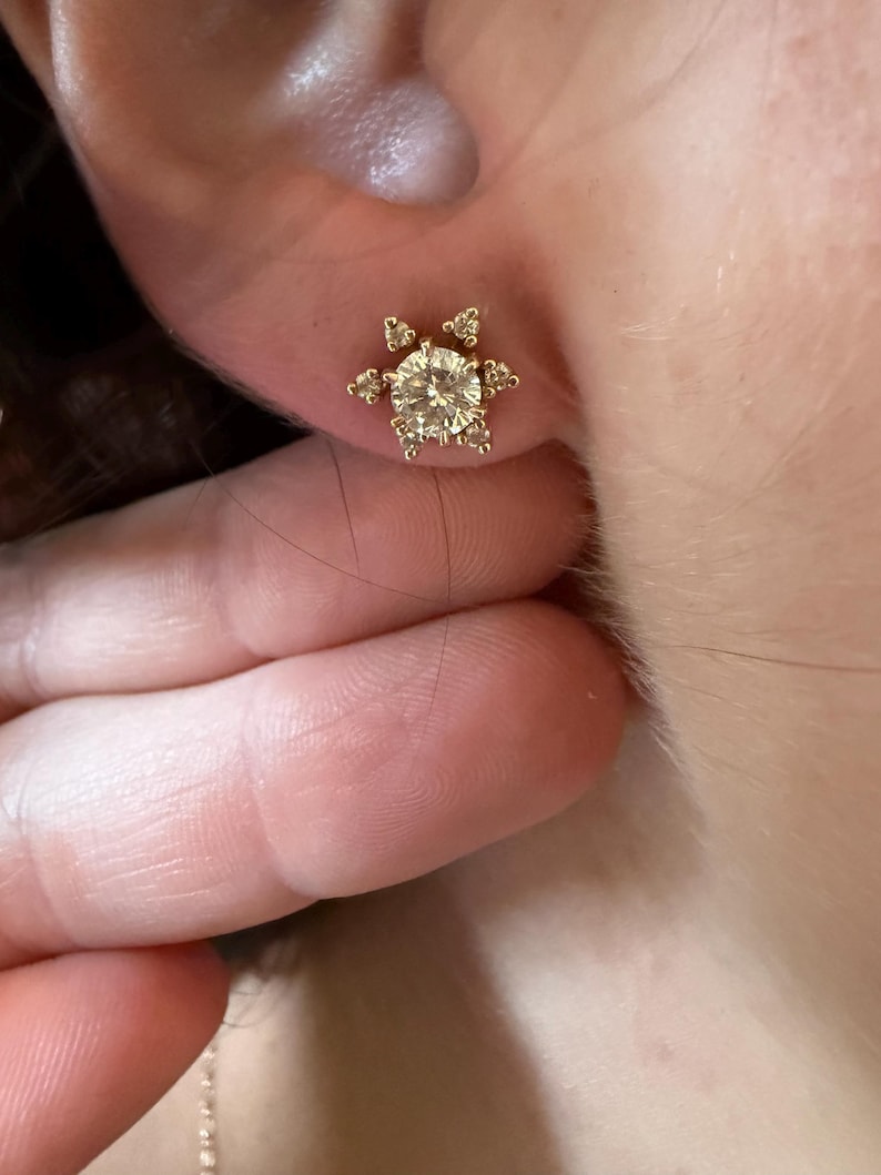 May include: A gold stud earring with a large, round, clear gemstone at the center, surrounded by smaller, clear gemstones in a starburst design. The earring is set in a gold metal setting.